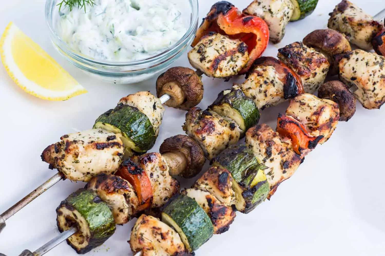three keto and low-carb lemon-oregano chicken kabobs on a plate with a side of tzatziki
