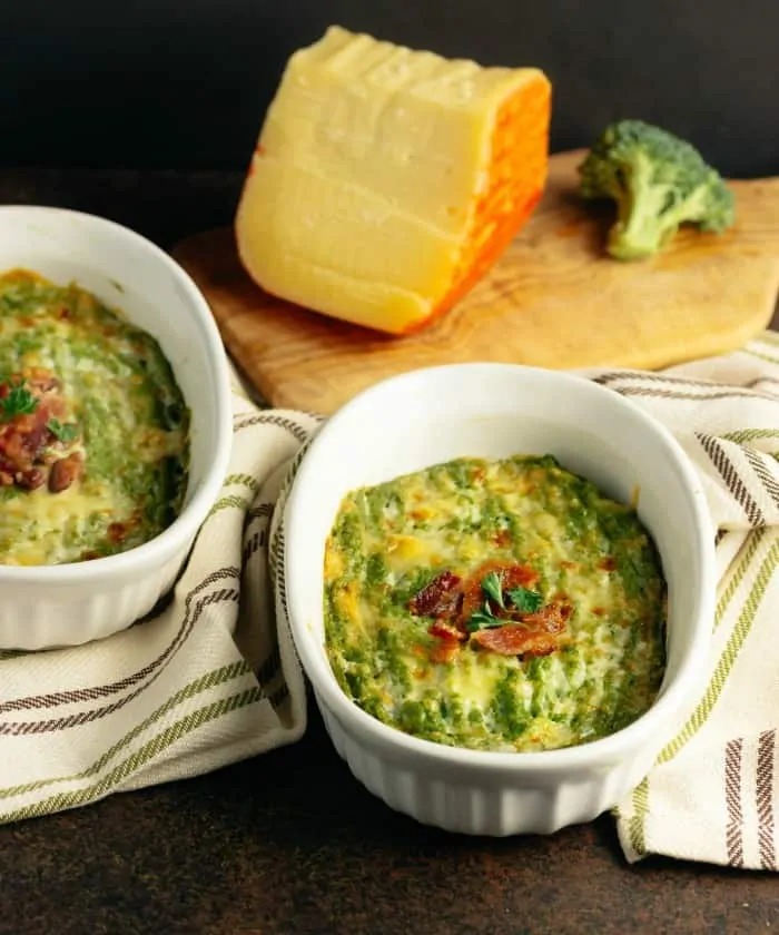 Two white bowls of Cheesy Broccoli Mash with Bacon and a wedge of cheese in the background.