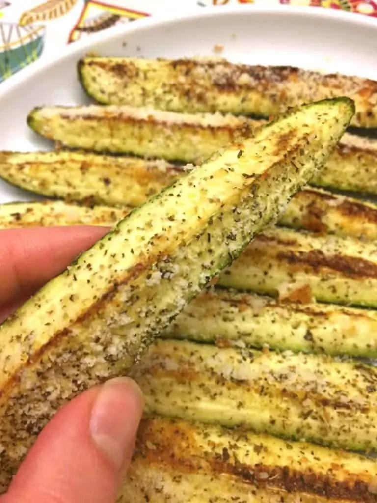 Fingers holding one Baked Parmesan-Garlic Zucchini spear above a plate of seven more. 