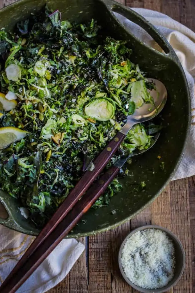 Kale and Brussels Sprouts Salad in a serving bowl with serving utensils and shredded Parmesan cheese.