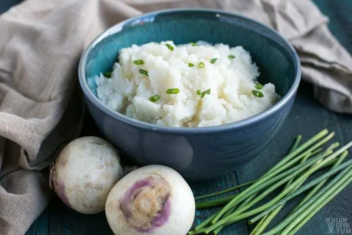 Mashed Turnips in a blue bowl next to raw turnips and chives.