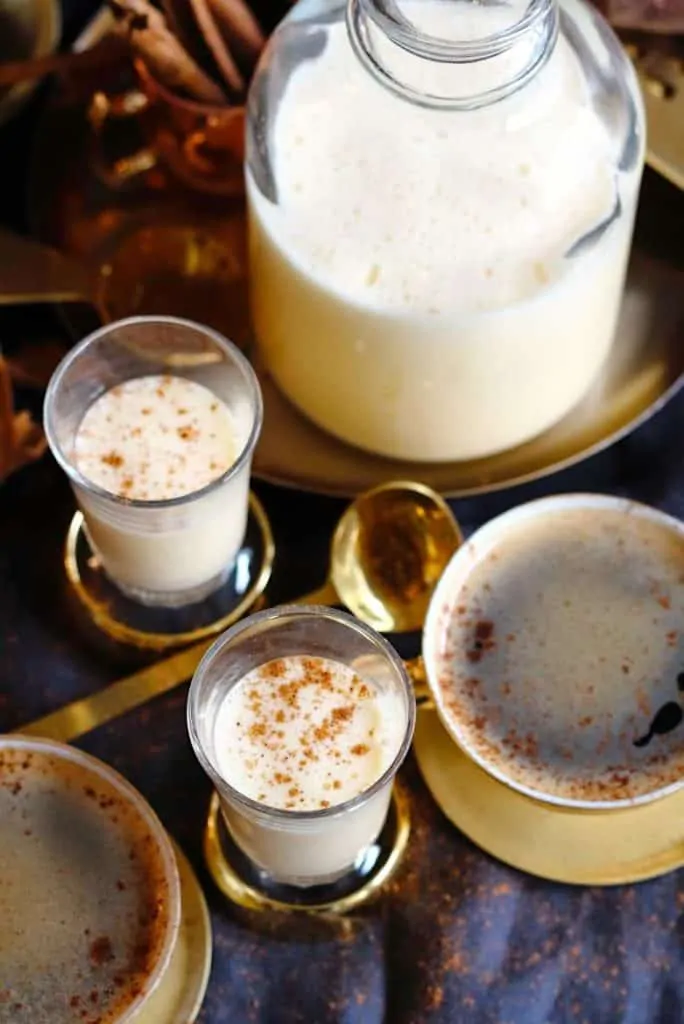 Two glasses of egg nog and two cups of egg nog coffee with a pitcher of egg nog in the background.