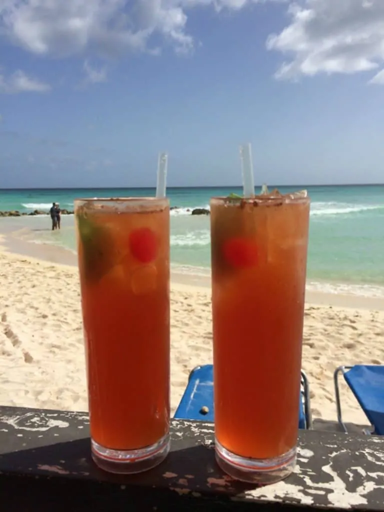 Two Barbados Rum Punch glasses with ice and straws on a beach in front of the water.