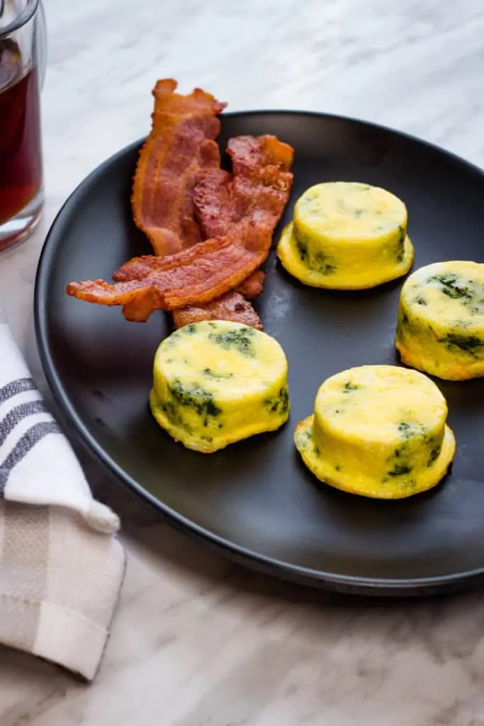 Four Keto Egg Bites with Spinach and Cheese on a black plate with bacon.