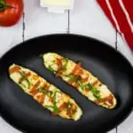 zucchini pizza boats on a black plate with a napkin and tomato in the background