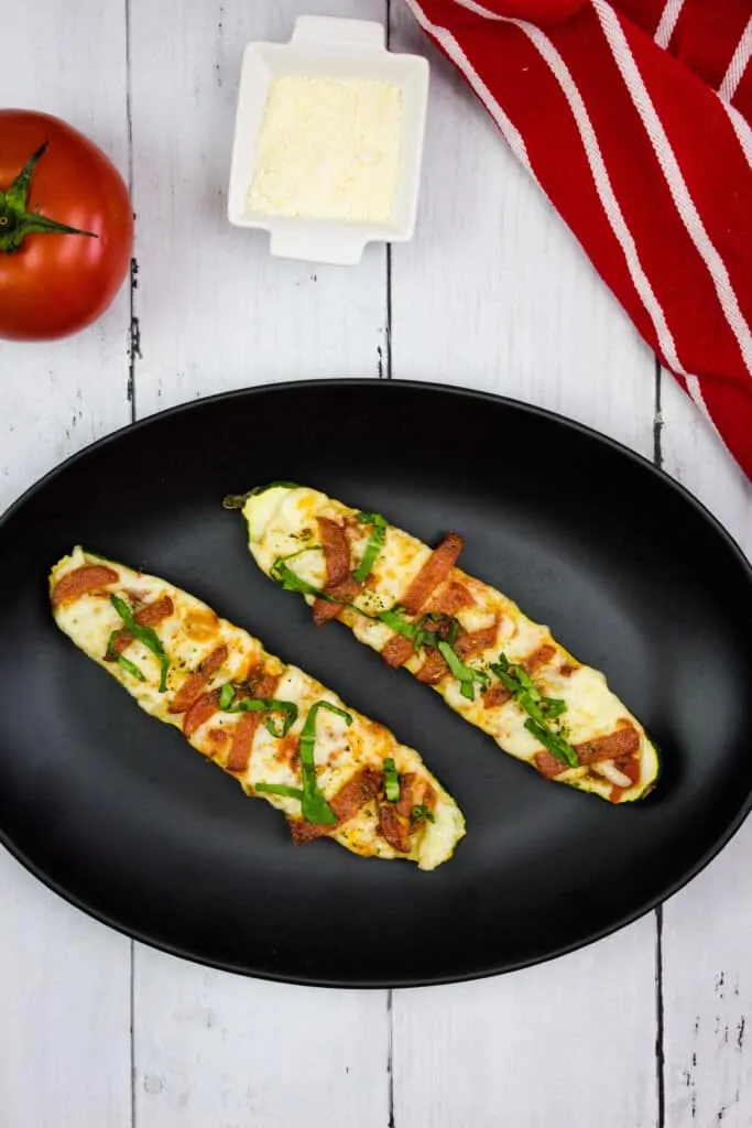 zucchini pizza boats on a black plate with a napkin and tomato in the background