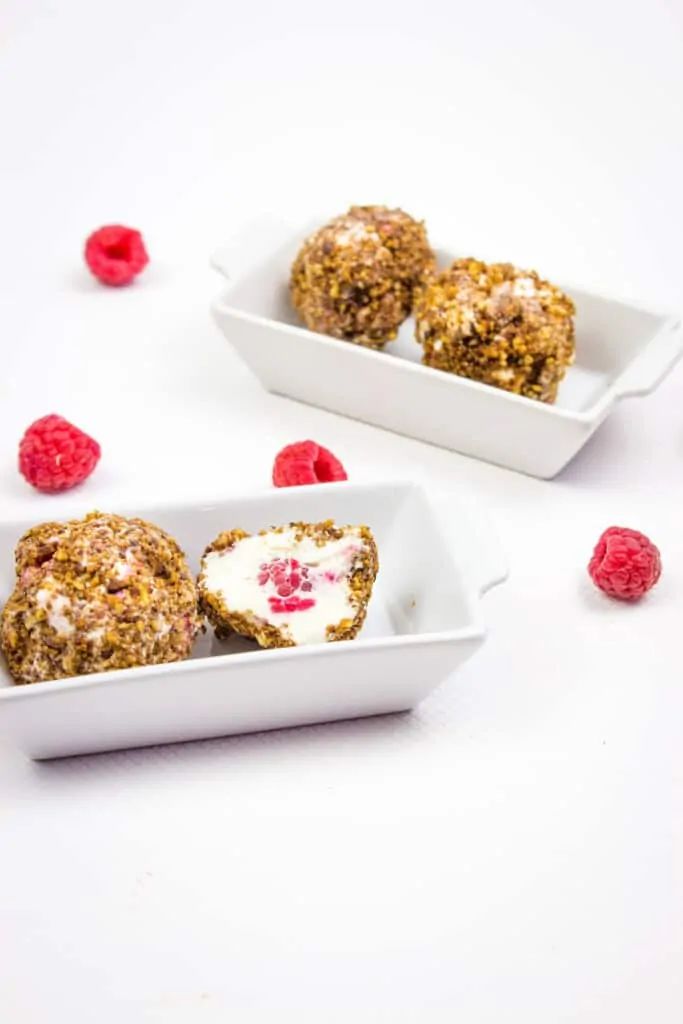 Raspberry frozen cheesecake balls in two small dishes. One is cut in half to show a raspberry in the center of the cheesecake ball.