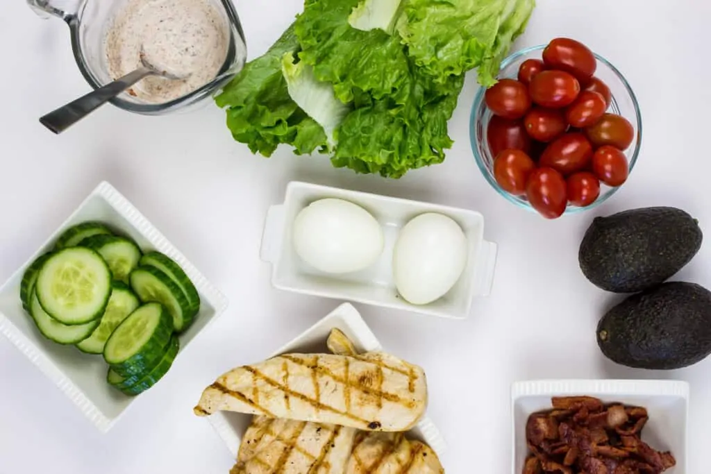 ingredients to make keto cobb salad (chicken, egg, tomatoes, lettuce, avocado, bacon, cucumbers, dressing)