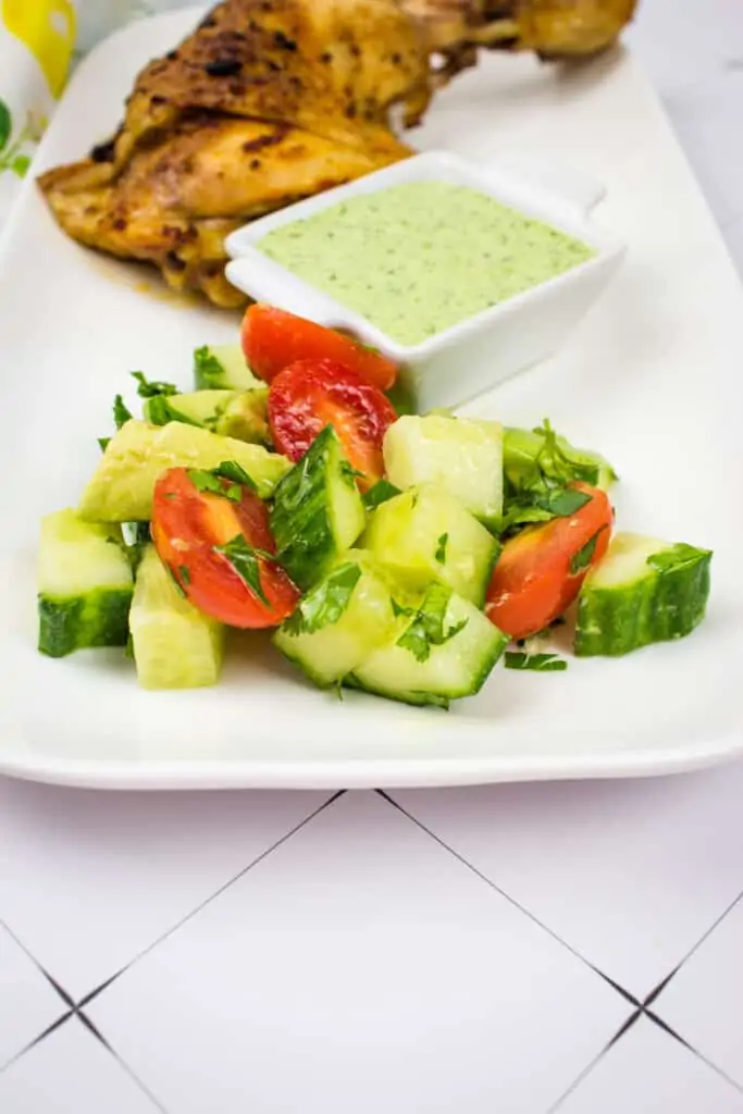 cucumber, tomato, avocado salad on a serving plate with peruvian chicken and green sauce in the background