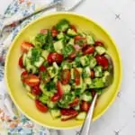 cucumber, tomato, avocado salad in a yellow serving bowl with a spoon and napkin in the background