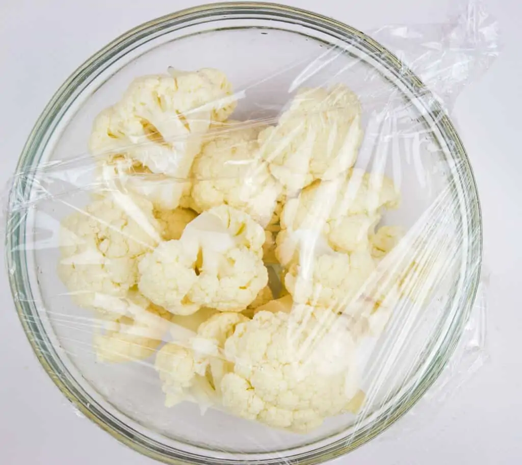 A bowl with cauliflower and water covered tightly with plastic wrap for steaming.