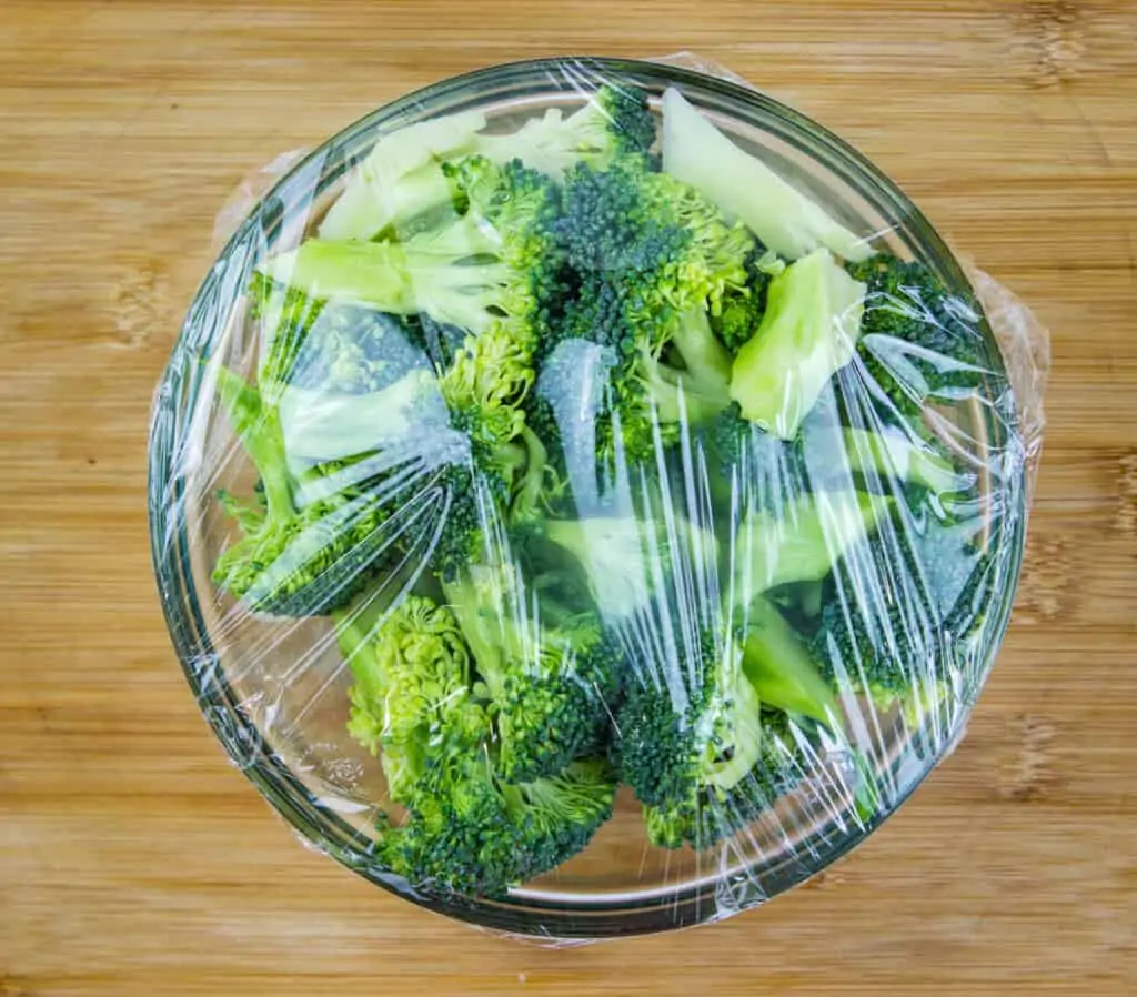 cut broccoli in a glass bowl with plastic wrap
