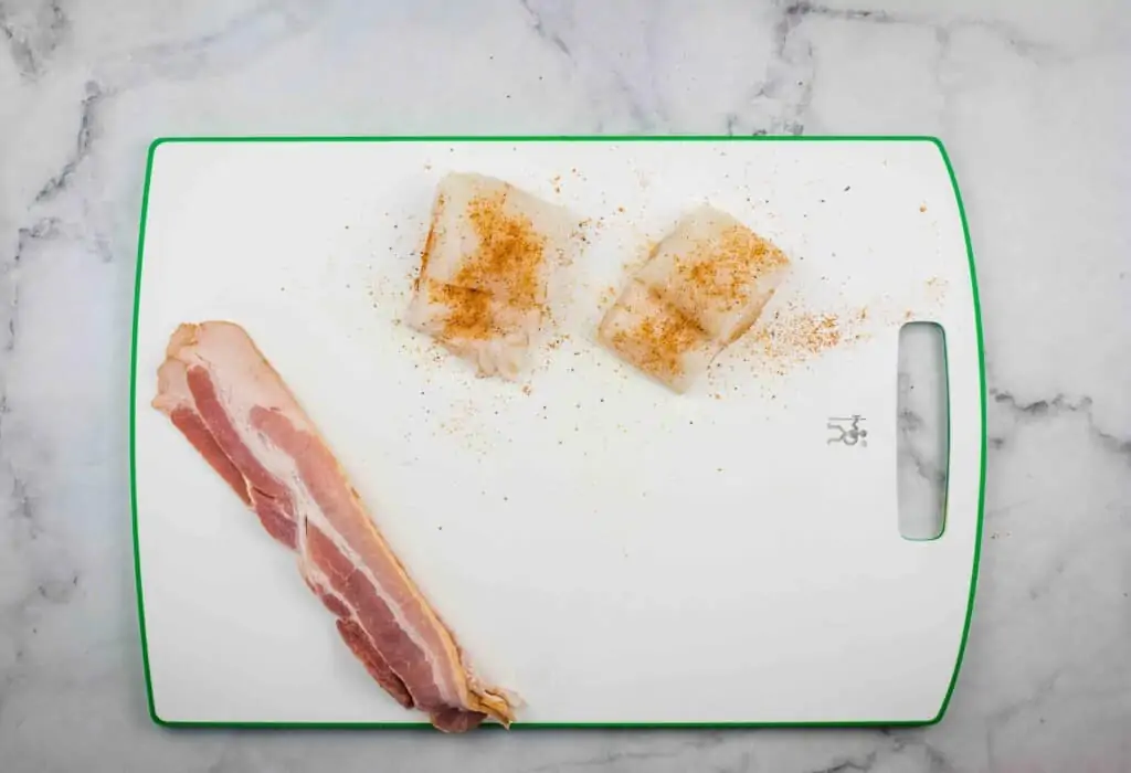 Seasoned cod and strips of bacon on a white cutting board.