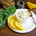 A closeup of keto tartar sauce in a white bowl with lemon and kale nearby.