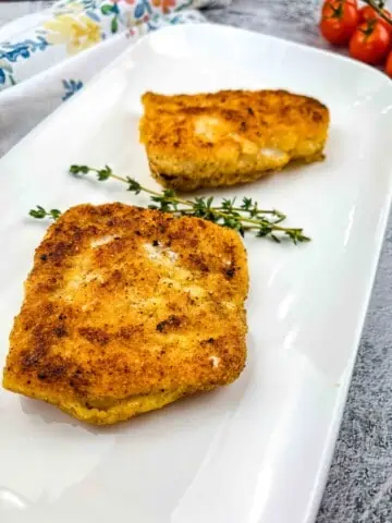 2 pieces of keto fried cod on a white plate with a sprig of thyme.