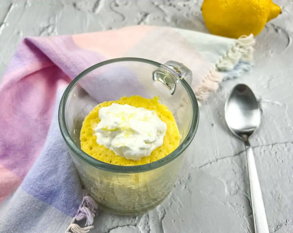 A horizontal shot of a keto lemon mug cake in a glass mug with a spoon to the side.