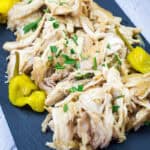 Low Carb Mississippi Chicken with chopped herbs and three yellow peppers on a black rectangular plate, placed on a gray surface with a floral cloth in the background.