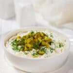 A creamy dill pickle dip garnished with finely chopped pickles and fresh dill, served in a white bowl on a matching white plate, set on a rustic white wooden table.