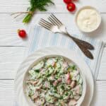 A bowl of fresh salad with sliced radishes, cucumbers, and herbs, dressed in a creamy white sauce, on a white plate with forks, radishes, fresh dill and yogurt on the surface.