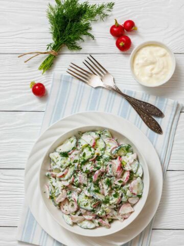 A bowl of fresh salad with sliced radishes, cucumbers, and herbs, dressed in a creamy white sauce, on a white plate with forks, radishes, fresh dill and yogurt on the surface.