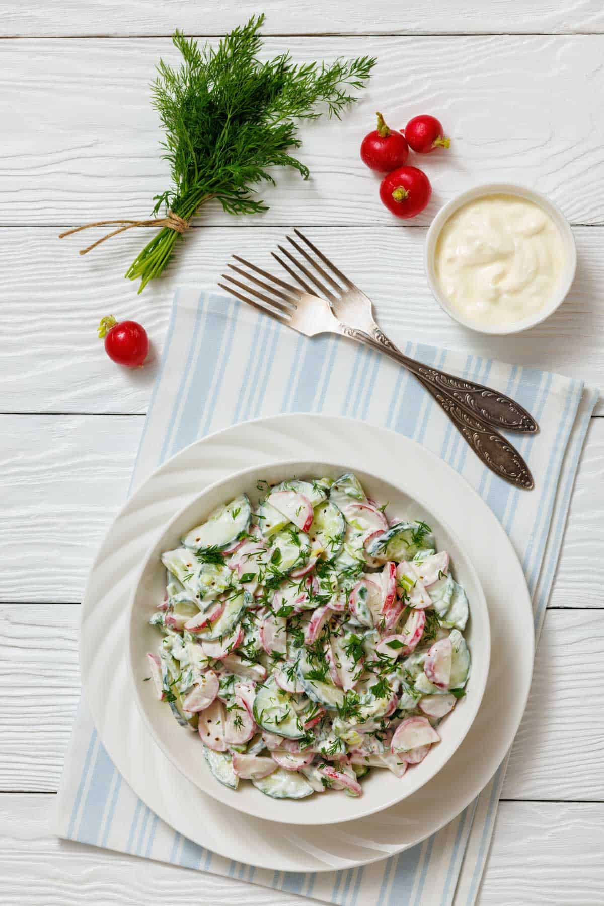 A bowl of fresh salad with sliced radishes, cucumbers, and herbs, dressed in a creamy white sauce, on a white plate with forks, radishes, fresh dill and yogurt on the surface.
