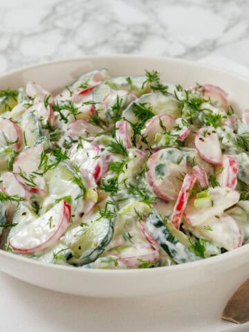 A bowl of cucumber and radish salad with dill and creamy dressing, placed on a white and blue napkin with a fork on the side.