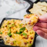 A hand holds a chip with Hot Reuben Dip from a skillet of the dip and cracker.