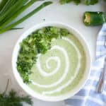 A white bowl of light green Low-Carb Cool & Creamy Cucumber Gazpacho, garnished with a swirl of cream, chopped cucumber, and herbs.