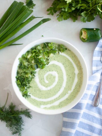 A white bowl of light green Low-Carb Cool & Creamy Cucumber Gazpacho, garnished with a swirl of cream, chopped cucumber, and herbs.
