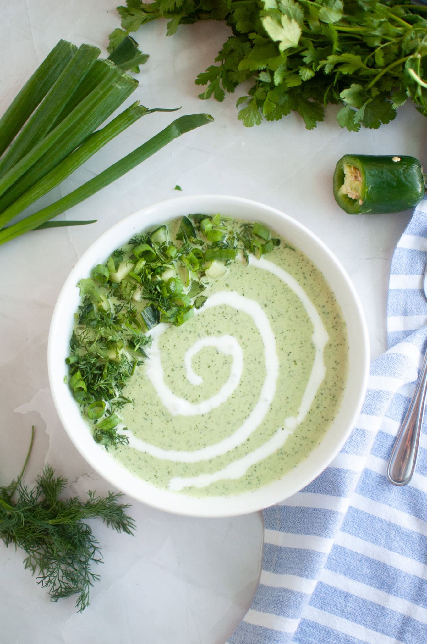 A white bowl of light green Low-Carb Cool & Creamy Cucumber Gazpacho, garnished with a swirl of cream, chopped cucumber, and herbs.