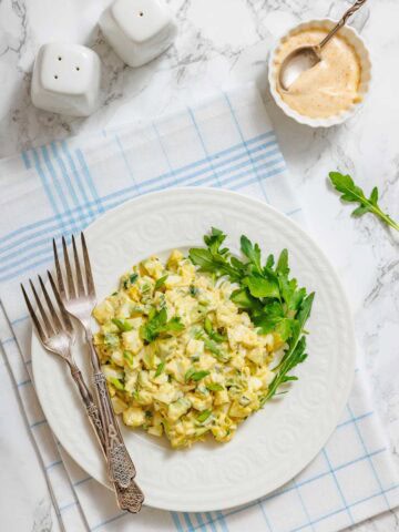 A white plate with egg salad, garnished with greens, is shown with forks, salt and pepper shakers, and a small bowl of sauce.