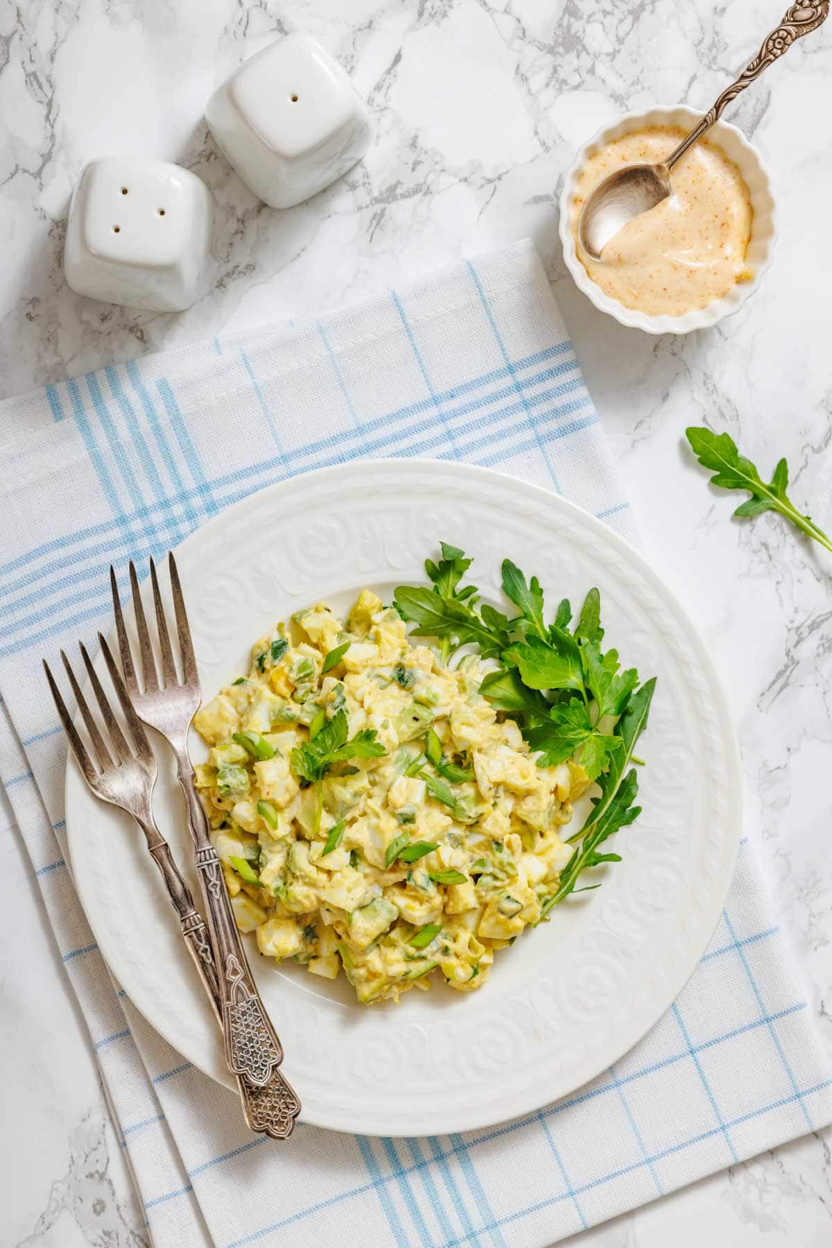 A white plate with egg salad, garnished with greens, is shown with forks, salt and pepper shakers, and a small bowl of sauce.