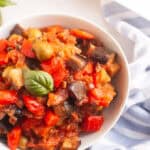 A white bowl filled with a colorful stew of eggplant, tomatoes, peppers, and olives, garnished with basil.