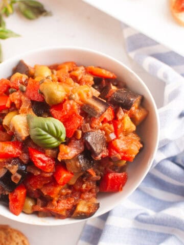 A white bowl filled with a colorful stew of eggplant, tomatoes, peppers, and olives, garnished with basil.