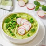 A white bowl of vibrant green soup, garnished with sliced radishes, fresh mint leaves, and green peas.