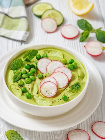 A white bowl of vibrant green soup, garnished with sliced radishes, fresh mint leaves, and green peas.