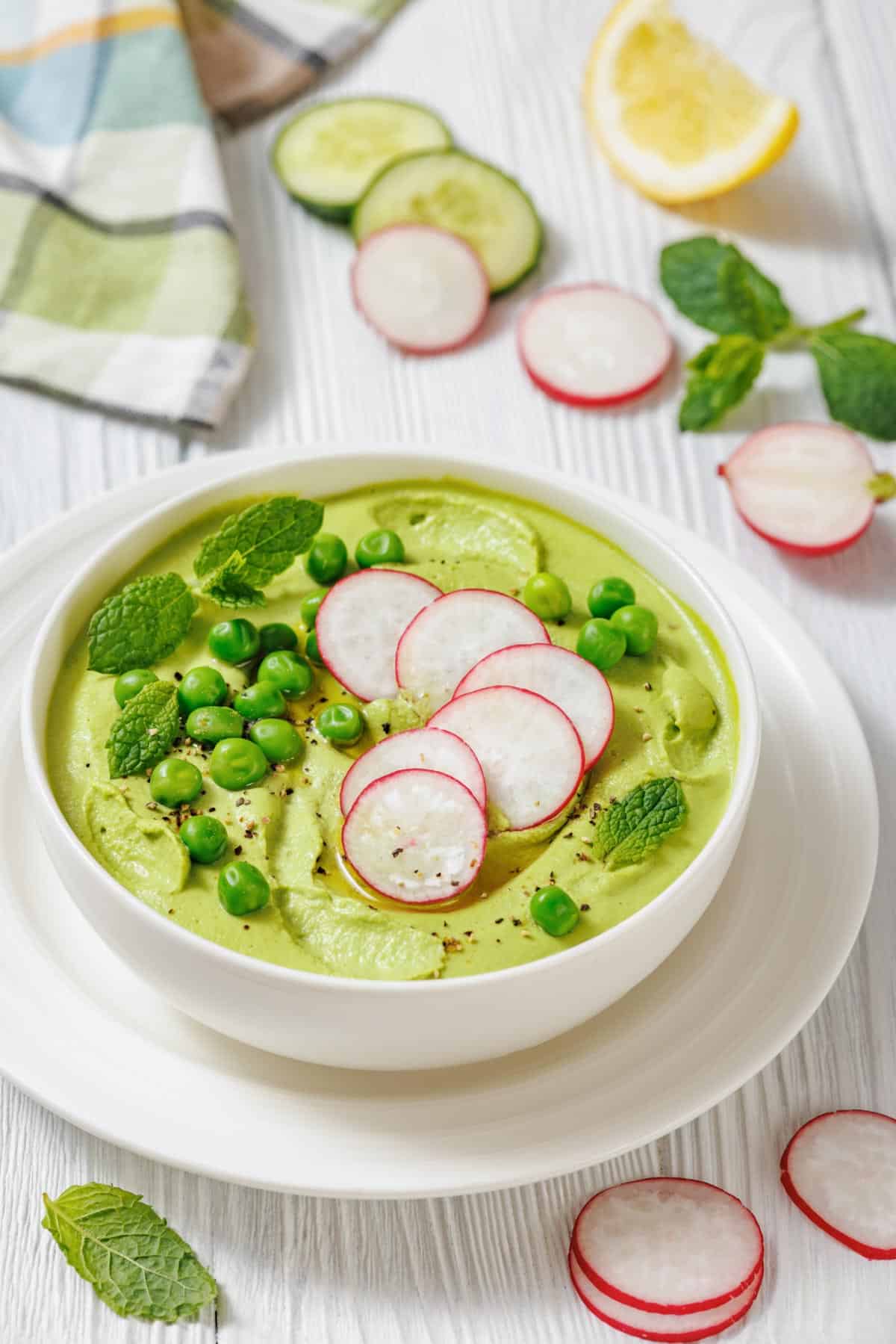 A white bowl of vibrant green soup, garnished with sliced radishes, fresh mint leaves, and green peas.