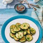 A white plate with sautéed zucchini slices, seasoned with garlic and herbs, is shown with a small bowl of chili flakes, a gravy boat of sauce, and salt and pepper shakers.