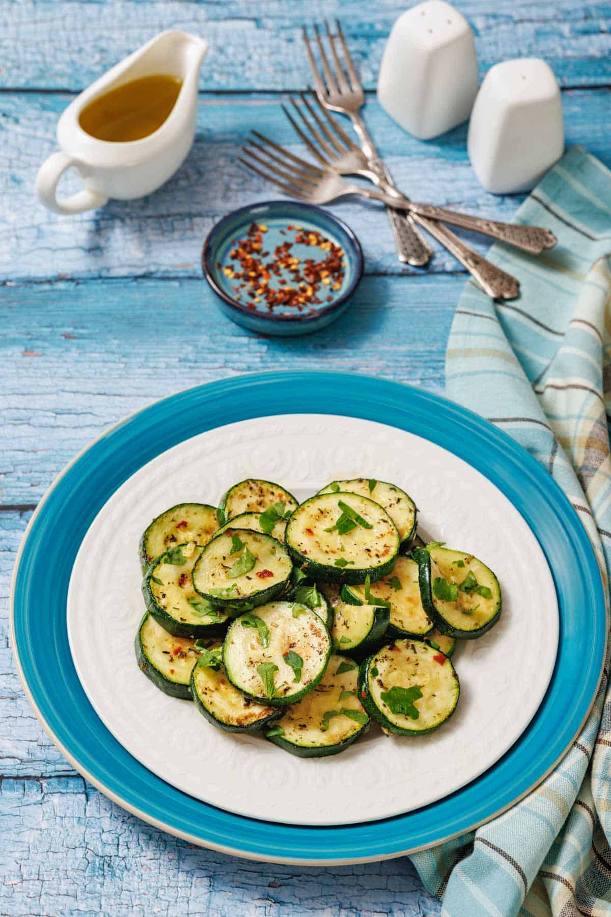 A white plate with sautéed zucchini slices, seasoned with garlic and herbs, is shown with a small bowl of chili flakes, a gravy boat of sauce, and salt and pepper shakers.