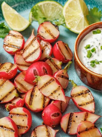 Grilled radishes with prominent char marks arranged on a turquoise plate, served with a creamy jalape&ntilde;o dip topped with chopped jalape&ntilde;os and cilantro, garnished with lime wedges and fresh cilantro leaves.