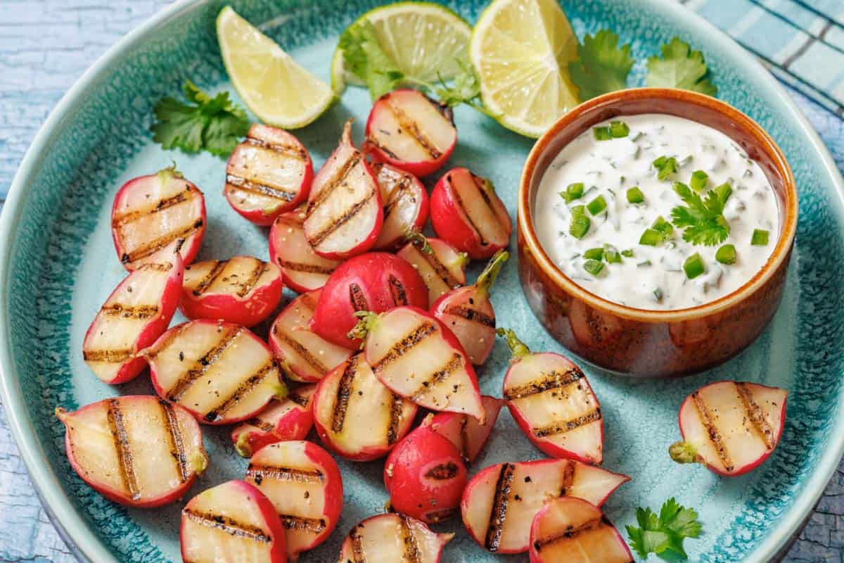 Grilled radishes with prominent char marks arranged on a turquoise plate, served with a creamy jalapeño dip topped with chopped jalapeños and cilantro, garnished with lime wedges and fresh cilantro leaves.