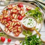 Char-grilled radish halves served on a round plate with a copper saucepan filled with creamy jalapeño dip, garnished with fresh cilantro, and surrounded by whole radishes and a colorful plaid cloth.