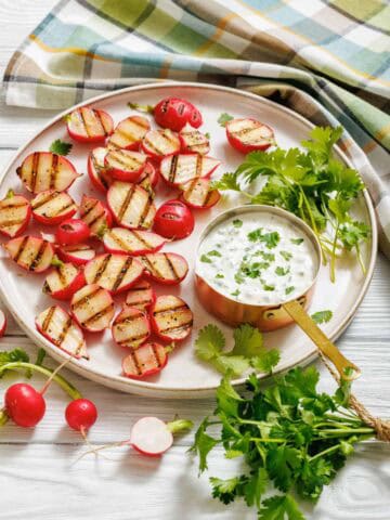 Char-grilled radish halves served on a round plate with a copper saucepan filled with creamy jalape&ntilde;o dip, garnished with fresh cilantro, and surrounded by whole radishes and a colorful plaid cloth.
