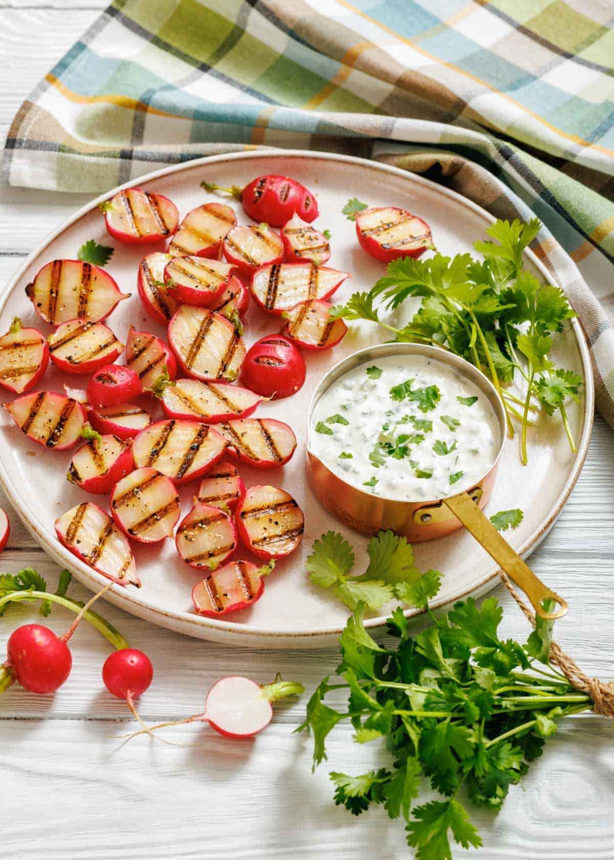 Char-grilled radish halves served on a round plate with a copper saucepan filled with creamy jalapeño dip, garnished with fresh cilantro, and surrounded by whole radishes and a colorful plaid cloth.
