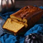A loaf of pumpkin pound cake with two slices cut sits on a glass plate atop a blue cloth, with decorative pumpkins nearby.
