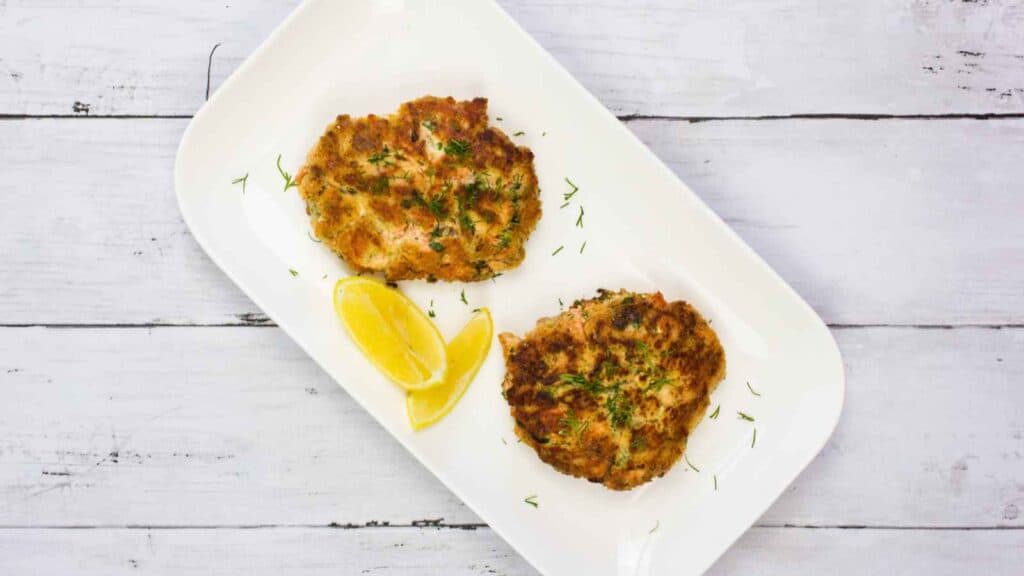 Top-down shot of two salmon patties arranged on a white rectangular plate, accompanied by lemon wedges.
