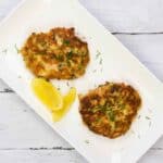Top-down shot of two salmon patties arranged on a white rectangular plate, accompanied by lemon wedges.