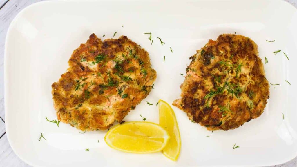 Two salmon patties arranged on a white rectangular plate, accompanied by lemon wedges.